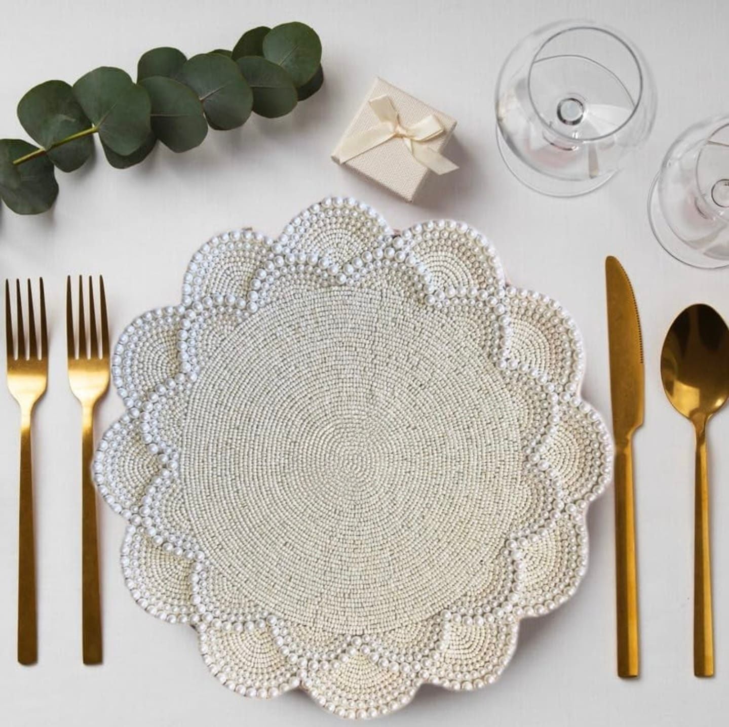 Round pearl-beaded white placemat with scalloped edges arranged on a light background, styled with gold cutlery, wine glasses, eucalyptus leaves, and a small gift box for an elegant table setting.