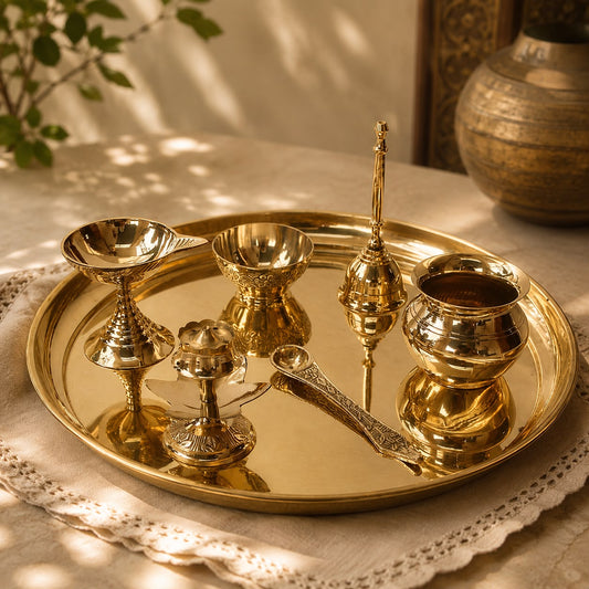 A polished brass or gold-toned metal serving set arranged on a round tray, including a small bowl, a tall spoon-like utensil, a small vessel with a lid, and a decorative spoon, photographed on a draped tablecloth with warm natural light and shadows.