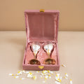 Two gold ladles in a pink velvet box on a beige surface with scattered flowers.