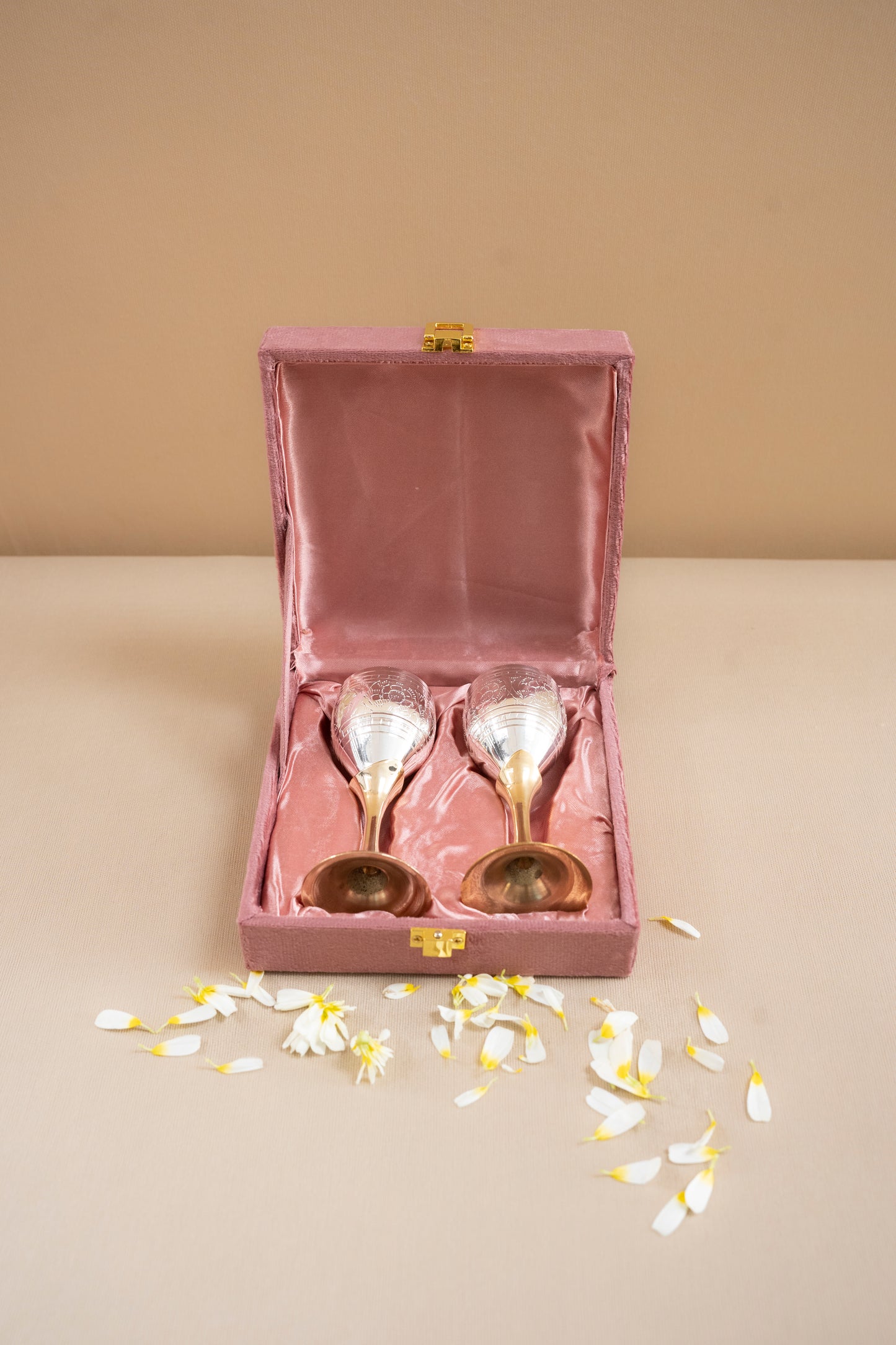 Two gold ladles in a pink velvet box on a beige surface with scattered flowers.