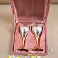 Two silver wine glasses in a pink velvet box with flower petals on a beige surface.