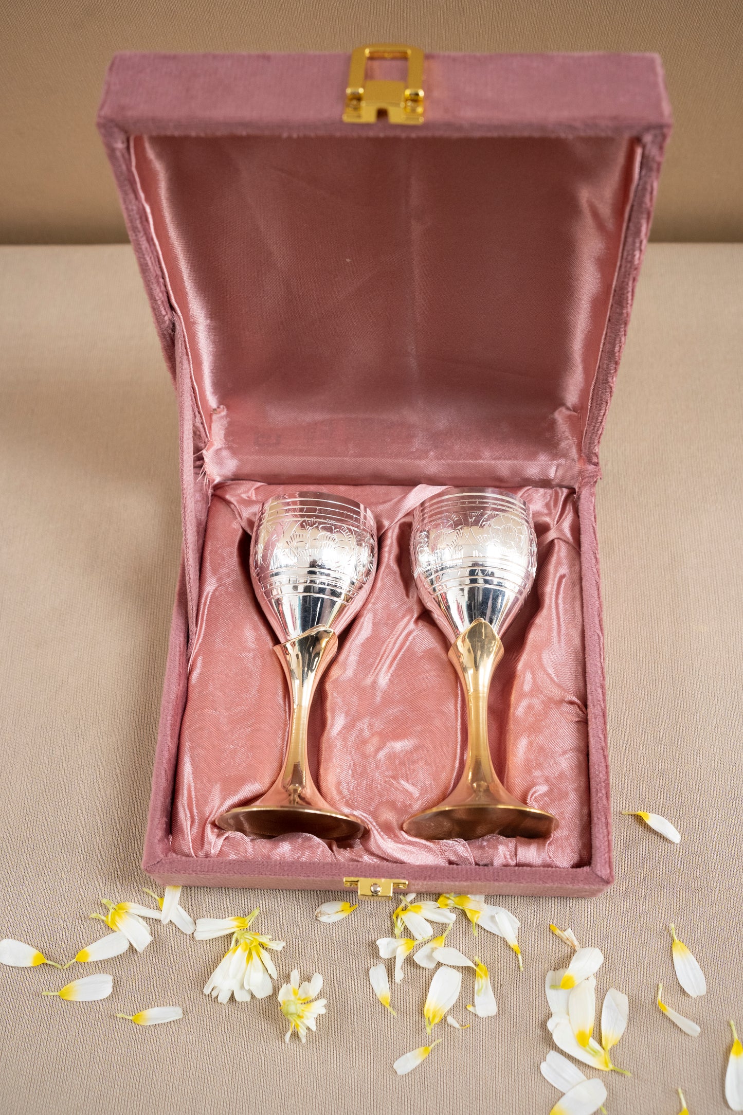 Two silver wine glasses in a pink velvet box with flower petals on a beige surface.