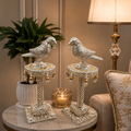 Decorative bird sculptures on stands with a lamp and plant in a room setting.