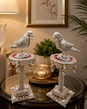Decorative parrot statues on stands with a candle and plant in the background