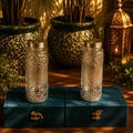 Two glass jars with silver lids on a blue velvet stand with potted plants and lanterns in the background.