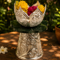 Decorative silver lotus-shaped container with flowers on a wooden surface with greenery in the background