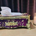 Decorative purple and gold tissue box with tissues on a table next to a lit candle.