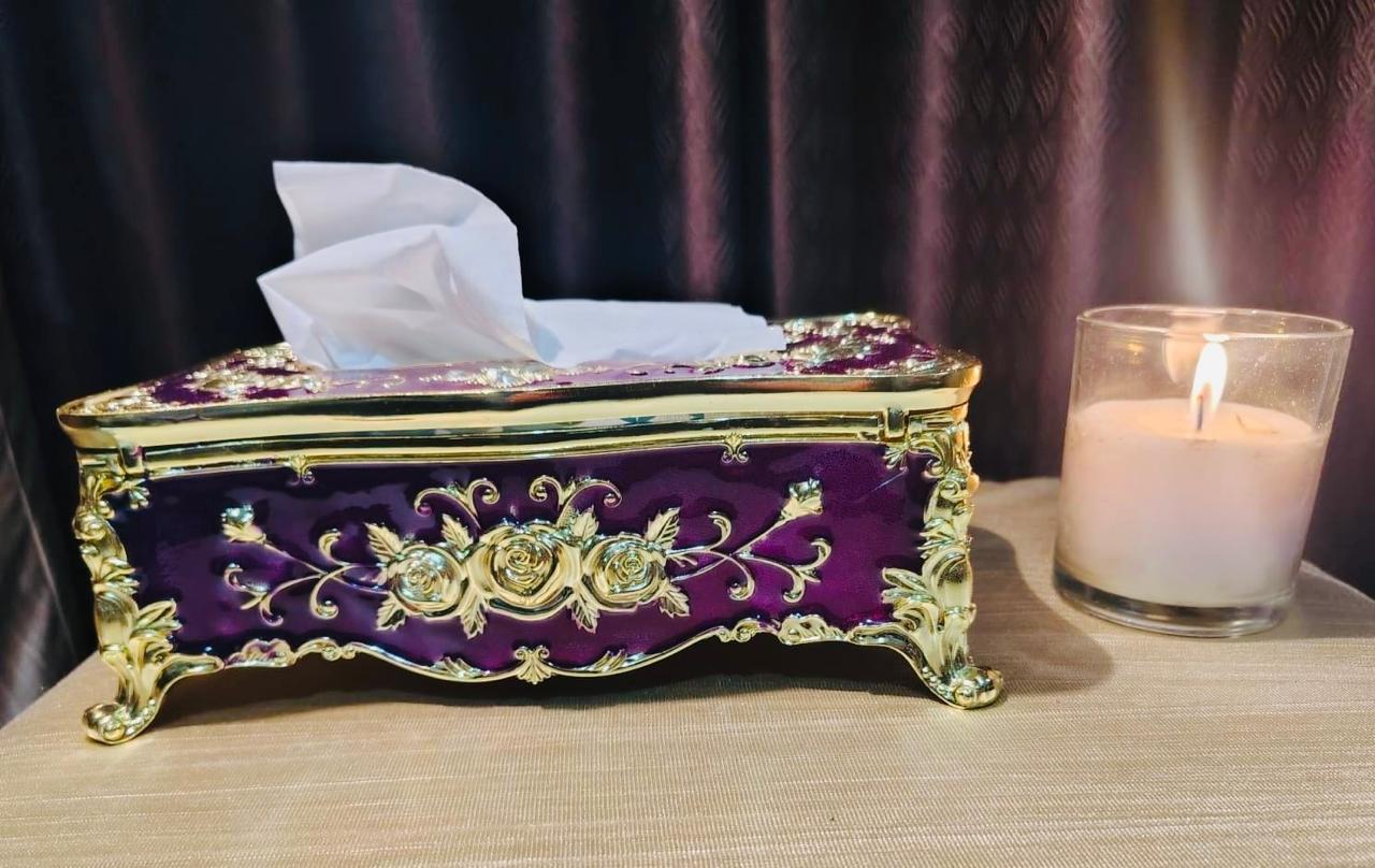 Decorative purple and gold tissue box with tissues on a table next to a lit candle.
