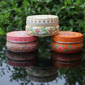 Three colorful decorative boxes stacked on a reflective surface with green foliage in the background