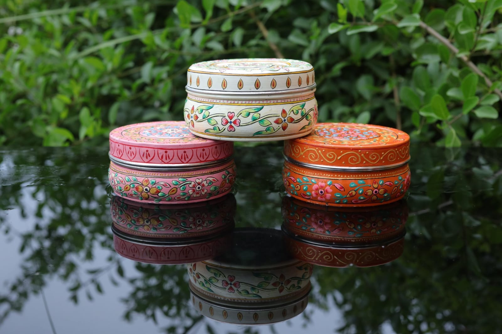 Three colorful decorative boxes stacked on a reflective surface with green foliage in the background