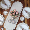 Elegant dining table setup featuring a pearl-beaded white table runner with scalloped edges, matching round placemats, white plates with folded napkins, candles, glassware, and a floral centerpiece on a wooden table.