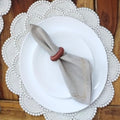 White dinner plate placed on a pearl-beaded round placemat with scalloped edges, styled with a folded beige napkin secured by a wooden napkin ring on a wooden table.