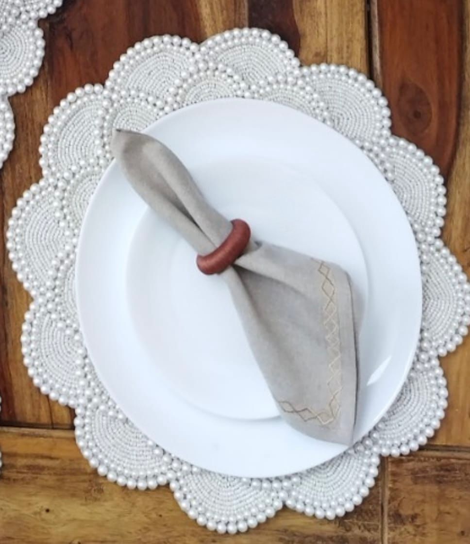 White dinner plate placed on a pearl-beaded round placemat with scalloped edges, styled with a folded beige napkin secured by a wooden napkin ring on a wooden table.