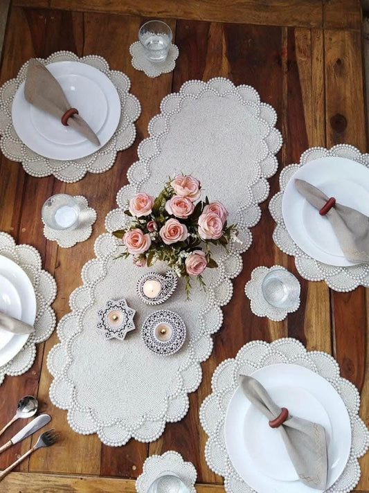 Elegant dining table setup featuring a pearl-beaded white table runner with scalloped edges, matching round placemats, white plates with folded napkins, candles, glassware, and a floral centerpiece on a wooden table.