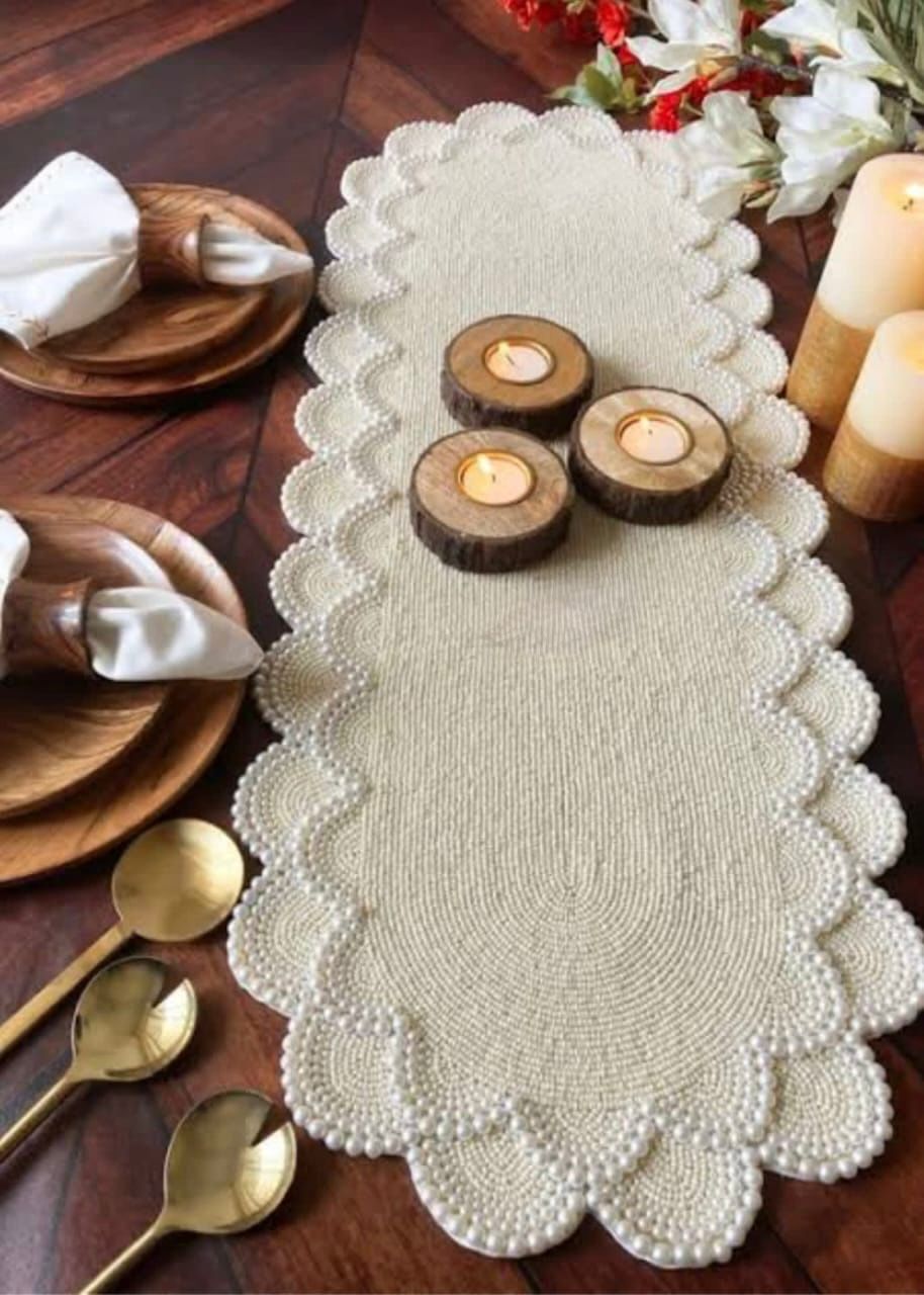 Pearl beaded white table runner with scalloped edges placed on a wooden dining table, styled with wooden candle holders, lit tea lights, gold cutlery, plates, and folded napkins for an elegant table setting.