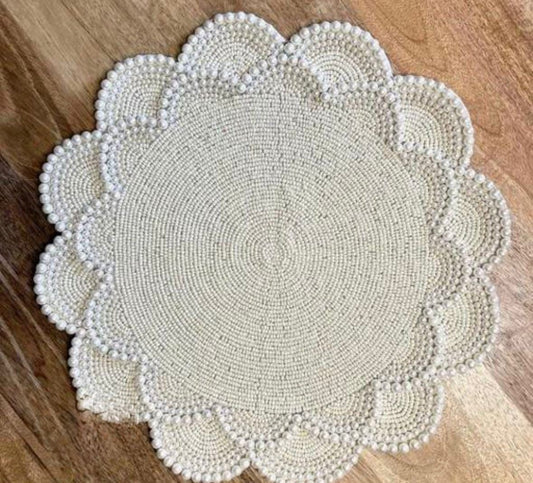Round pearl-beaded white placemat with intricate concentric patterns and scalloped edges, placed on a wooden surface.