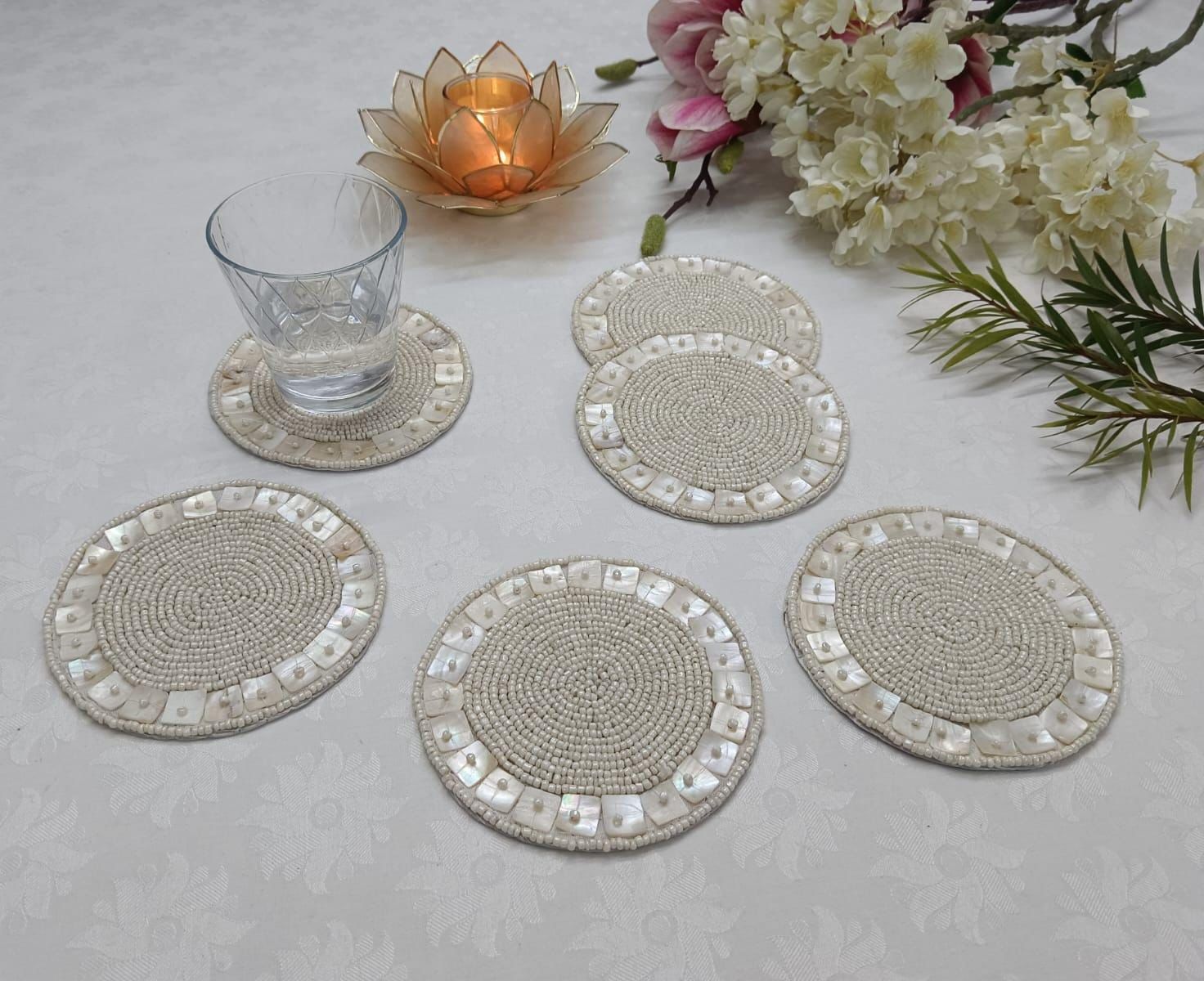 Set of round beaded coasters on a light surface with a glass, candle, and flowers.