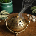 Bronze incense burner with smoke on a wooden surface with plants in the background
