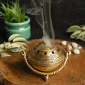 Bronze incense burner with smoke on a wooden surface with plants in the background