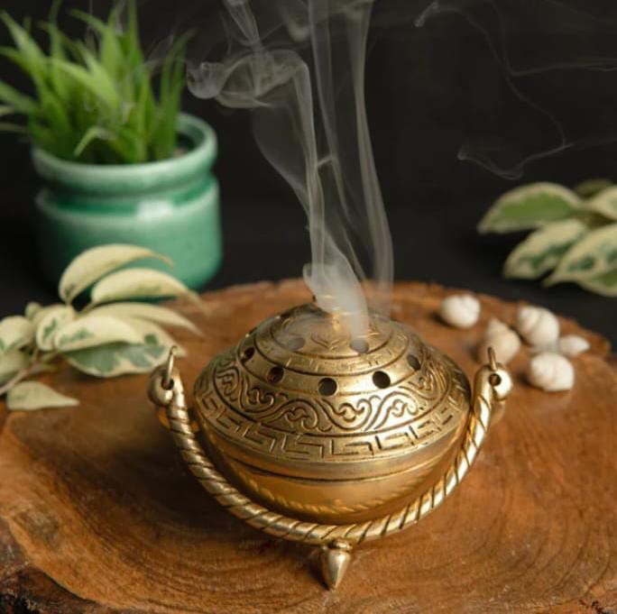 Bronze incense burner with smoke on a wooden surface with plants in the background