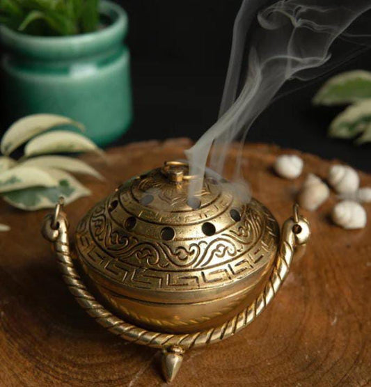 Bronze incense burner with smoke on a wooden surface with plants in the background