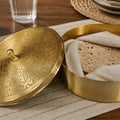 Brass bread box with a lid on a wooden table with a glass of water and plant in the background.