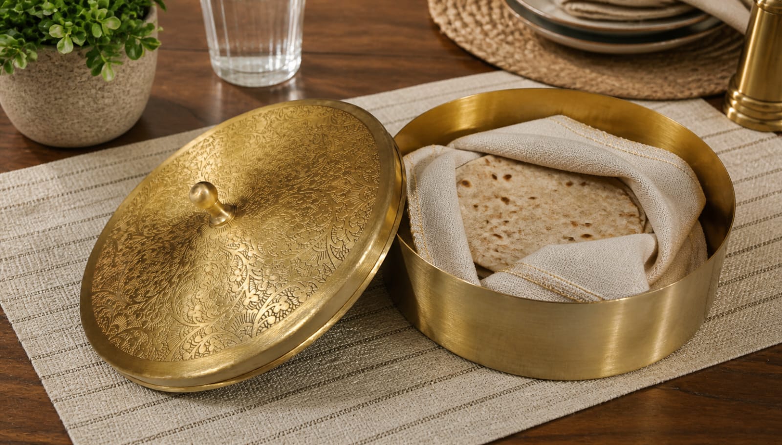 Brass bread box with a lid on a wooden table with a glass of water and plant in the background.