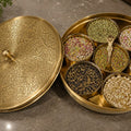 Decorative brass box with lid open, revealing assorted grains and seeds on a stone surface.