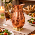 A hammered copper water pitcher with a polished brass handle and base, set on a dining table surrounded by salads, bread, and lit candles.