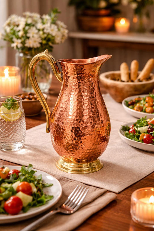A hammered copper water pitcher with a polished brass handle and base, set on a dining table surrounded by salads, bread, and lit candles.