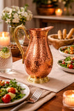 A hammered copper water pitcher with a polished brass handle and base, set on a dining table surrounded by salads, bread, and lit candles.