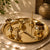A polished brass or gold-toned metal serving set arranged on a round tray, including a small bowl, a tall spoon-like utensil, a small vessel with a lid, and a decorative spoon, photographed on a draped tablecloth with warm natural light and shadows.