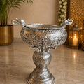 Decorative silver urn on a marble surface with a plant and candle in the background