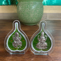 Two silver figurines on green leaves in clear stands on a wooden surface with a green pot in the background.