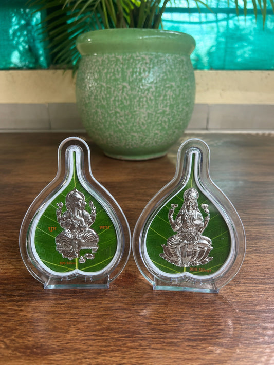 Two silver figurines on green leaves in clear stands on a wooden surface with a green pot in the background.