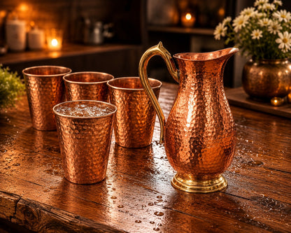 A hammered copper pitcher with a curved handle and four matching copper cups arranged on a rustic wooden table in a warmly lit indoor setting.