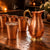 A hammered copper pitcher with a curved handle and four matching copper cups arranged on a rustic wooden table in a warmly lit indoor setting.