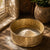 An ornate gold metal bowl with embossed floral patterns sits on a wooden table in warm sunlight, with a small pile of white powder inside. Dried flowers, a ceramic planter, and a window frame are visible in the softly lit background.