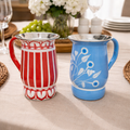 Two ceramic pitchers, one red and white striped, the other blue with floral design, on a table with a floral arrangement in the background.