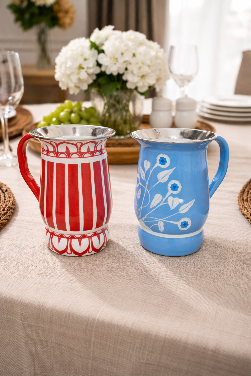 Two ceramic pitchers, one red and white striped, the other blue with floral design, on a table with a floral arrangement in the background.